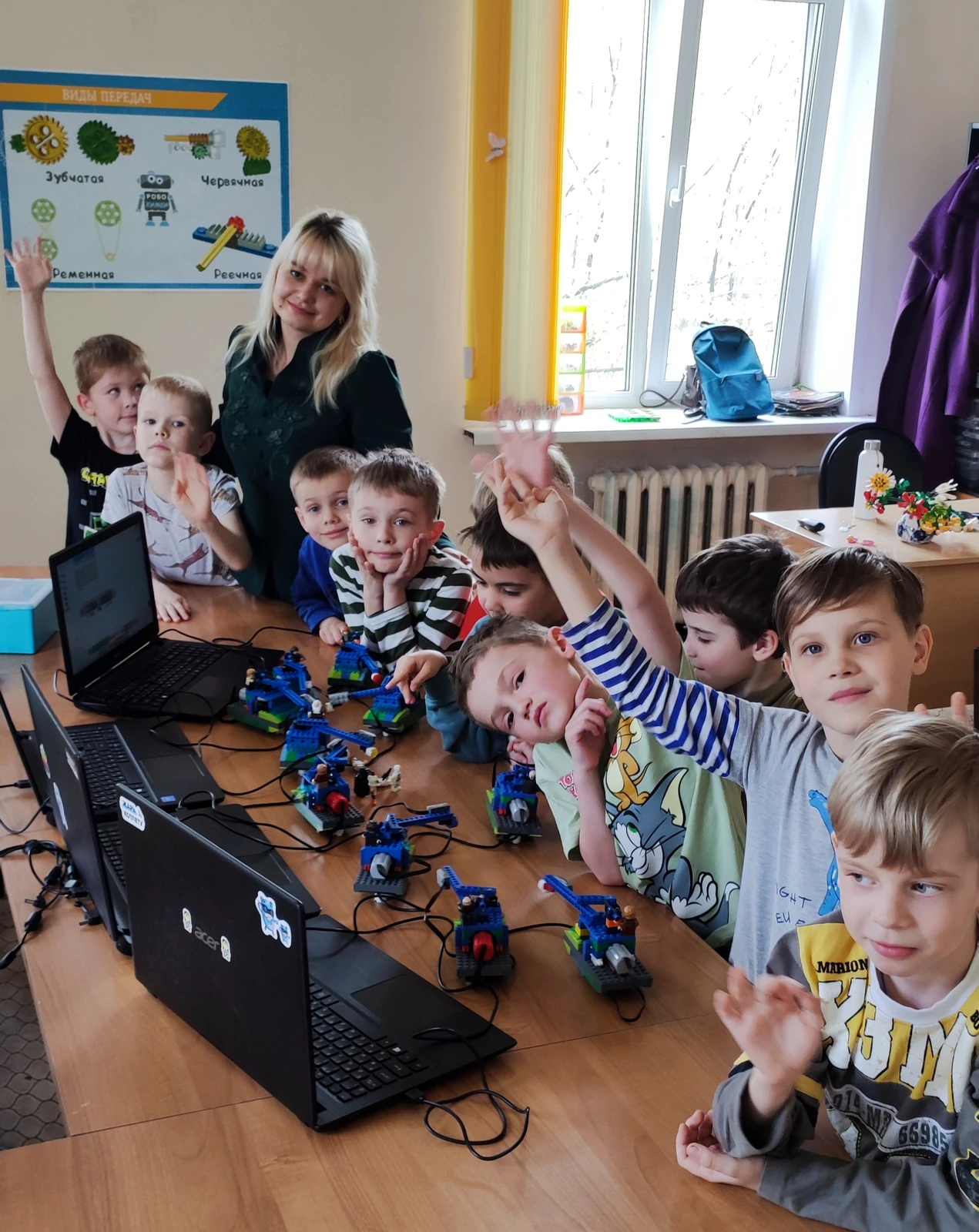 Преподаватель и большая группа учеников вокруг стола с ноутбуками и роботами