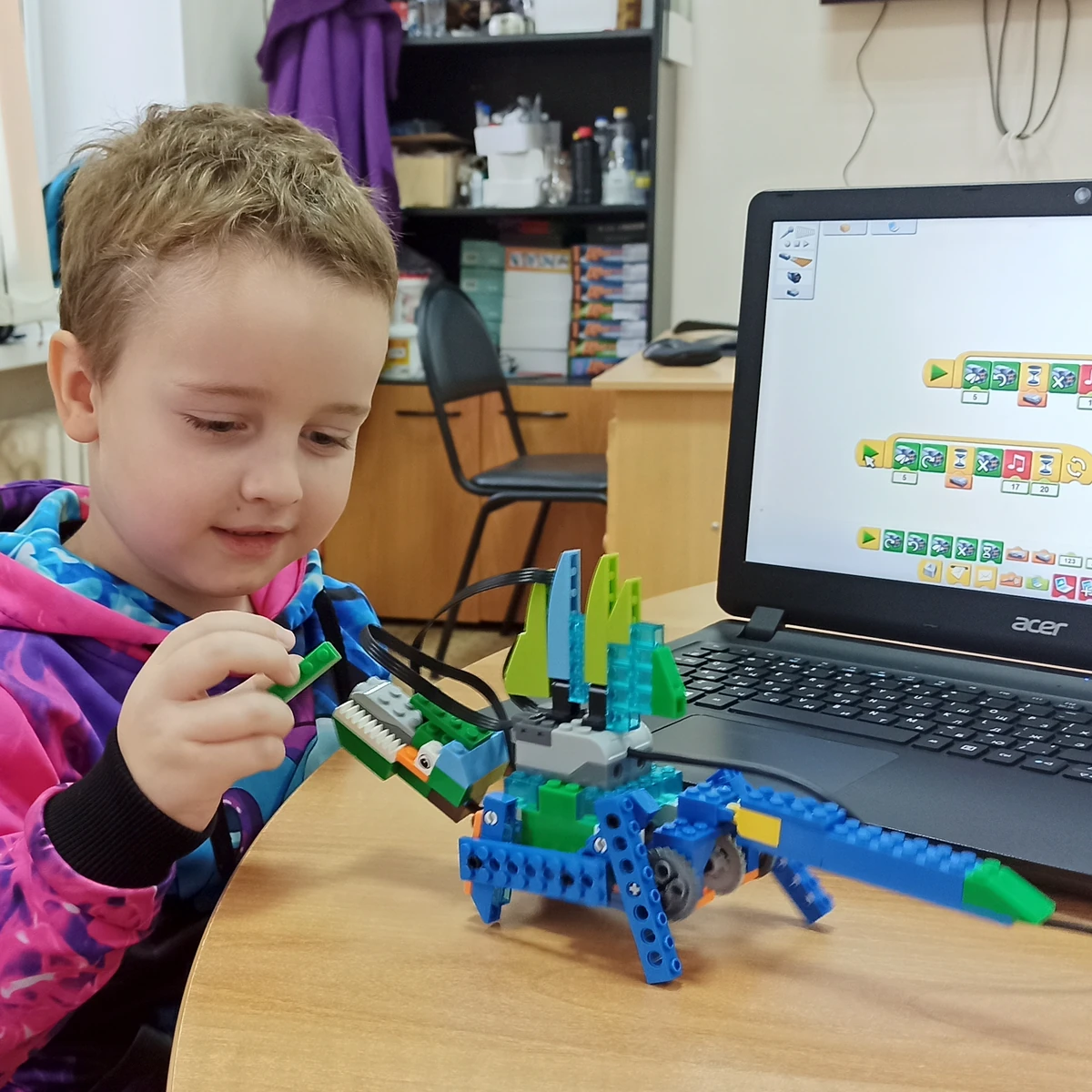 Ученик в яркой толстовке собирает модель динозавра LEGO у ноутбука